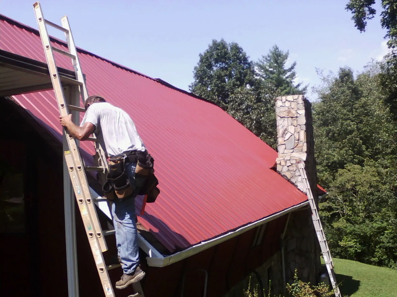 Roofer on a ladder beside a red metal roof and stone chimney — Soffit & Fascia Repair by Roofing Brothers in Webster